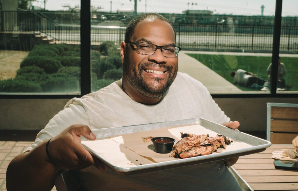 Quincy James Freeman smiling while holding a piece of brisket, with a tray of barbecue on the table