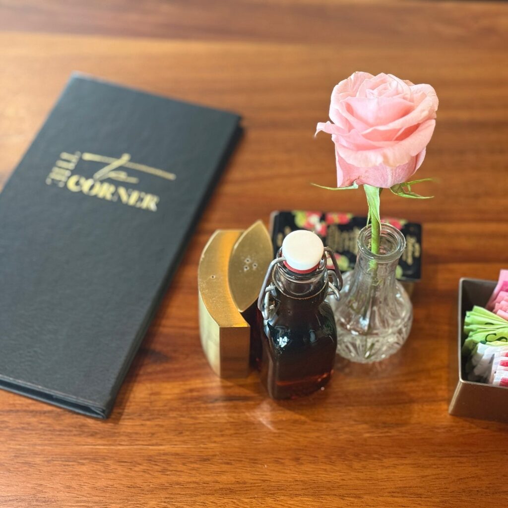 A table setting at The Corner with a menu book, a bottle of syrup, and a pink rose