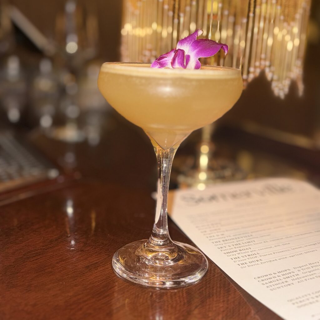 The D.W. cocktail at Somerville's bar, served in a coupe glass and garnished with a purple orchid, beneath a gold beaded chandelier.