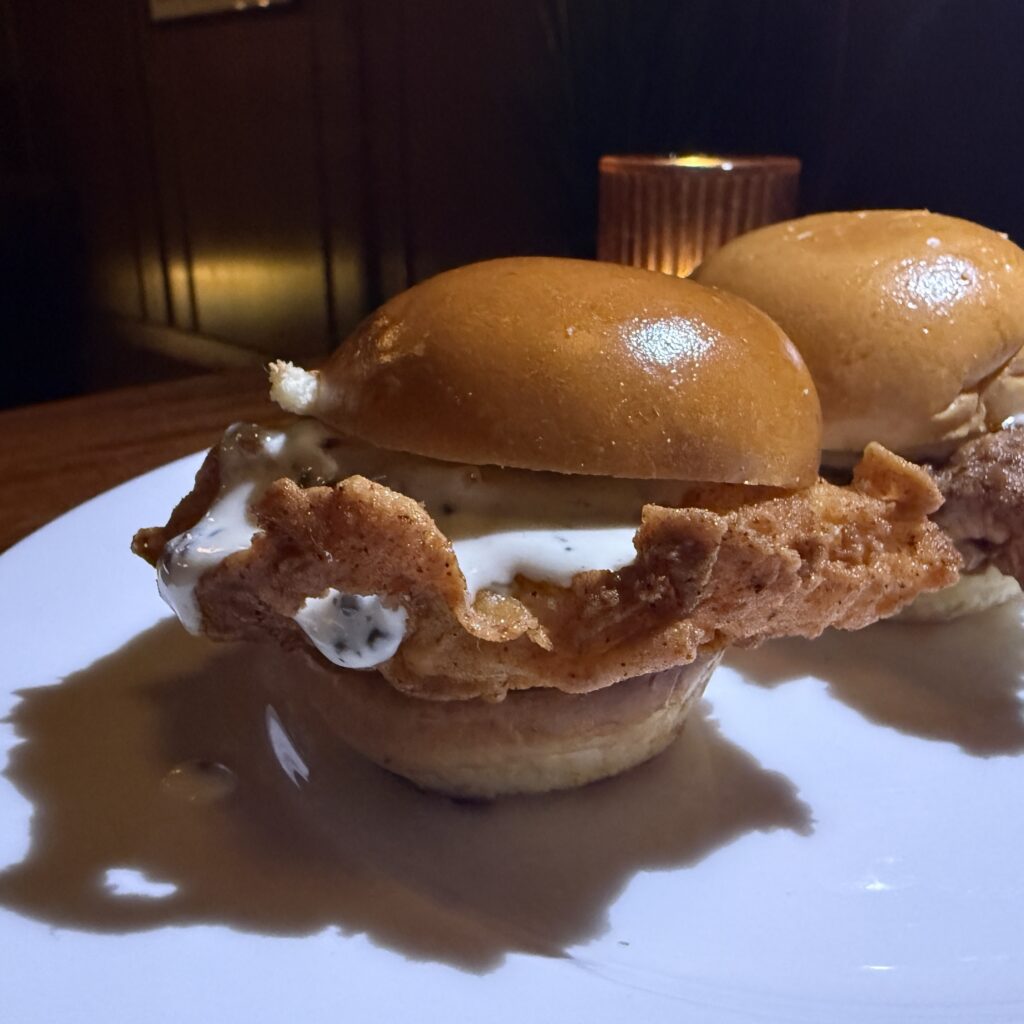 A bitten fried chicken and caviar slider showing the crispy fried chicken, caviar crème fraîche, and bread-and-butter pickle inside a brioche bun.