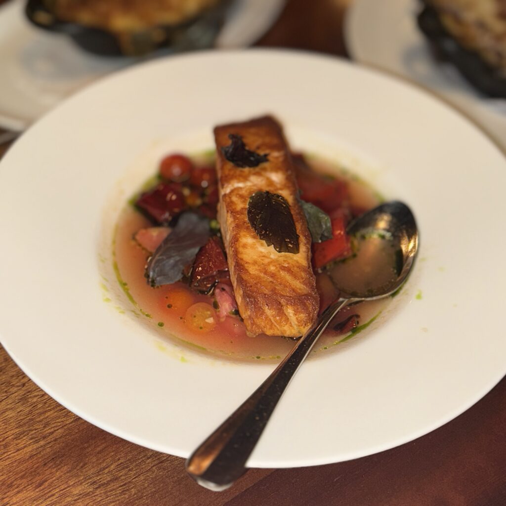 A seared Scottish salmon fillet plated over heirloom tomatoes and cherry tomatoes in a light tomato broth, garnished with purple basil leaves.