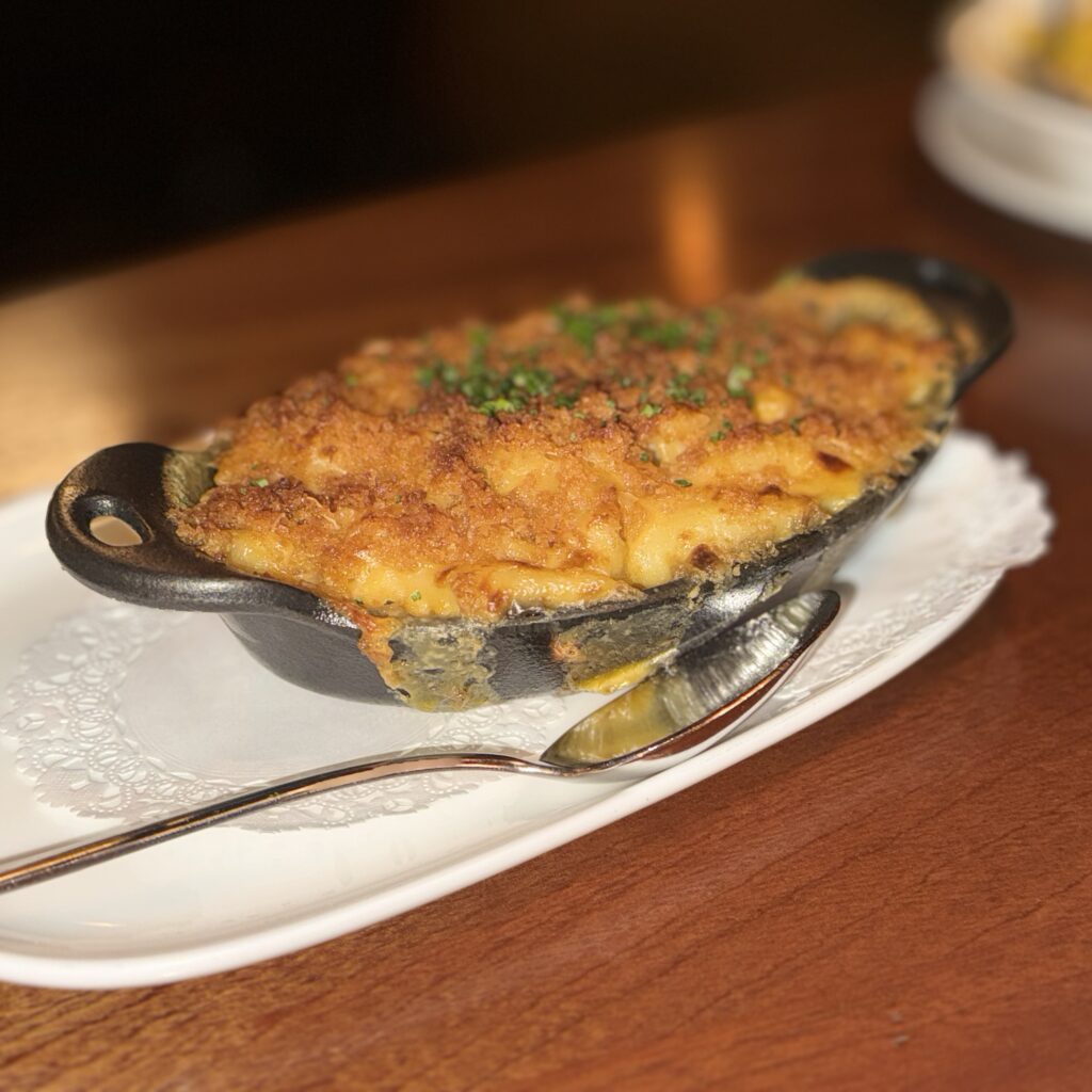 Close-up of Somerville's baked macaroni and cheese served in a mini cast-iron skillet, with a golden breadcrumb crust and chopped chives on top, alongside a pickle spear.