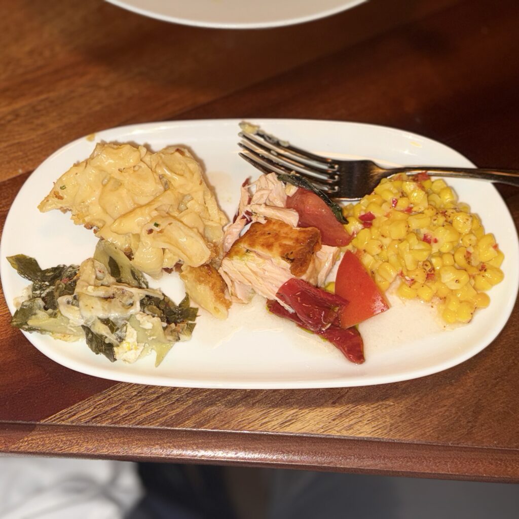 Overhead shot of a shared plate with a scoop of baked macaroni and cheese, a portion of collard green lasagna, a piece of seared Scottish salmon over tomatoes, and a side of buttered corn with pickled fresno chiles, with a fork resting across the plate.